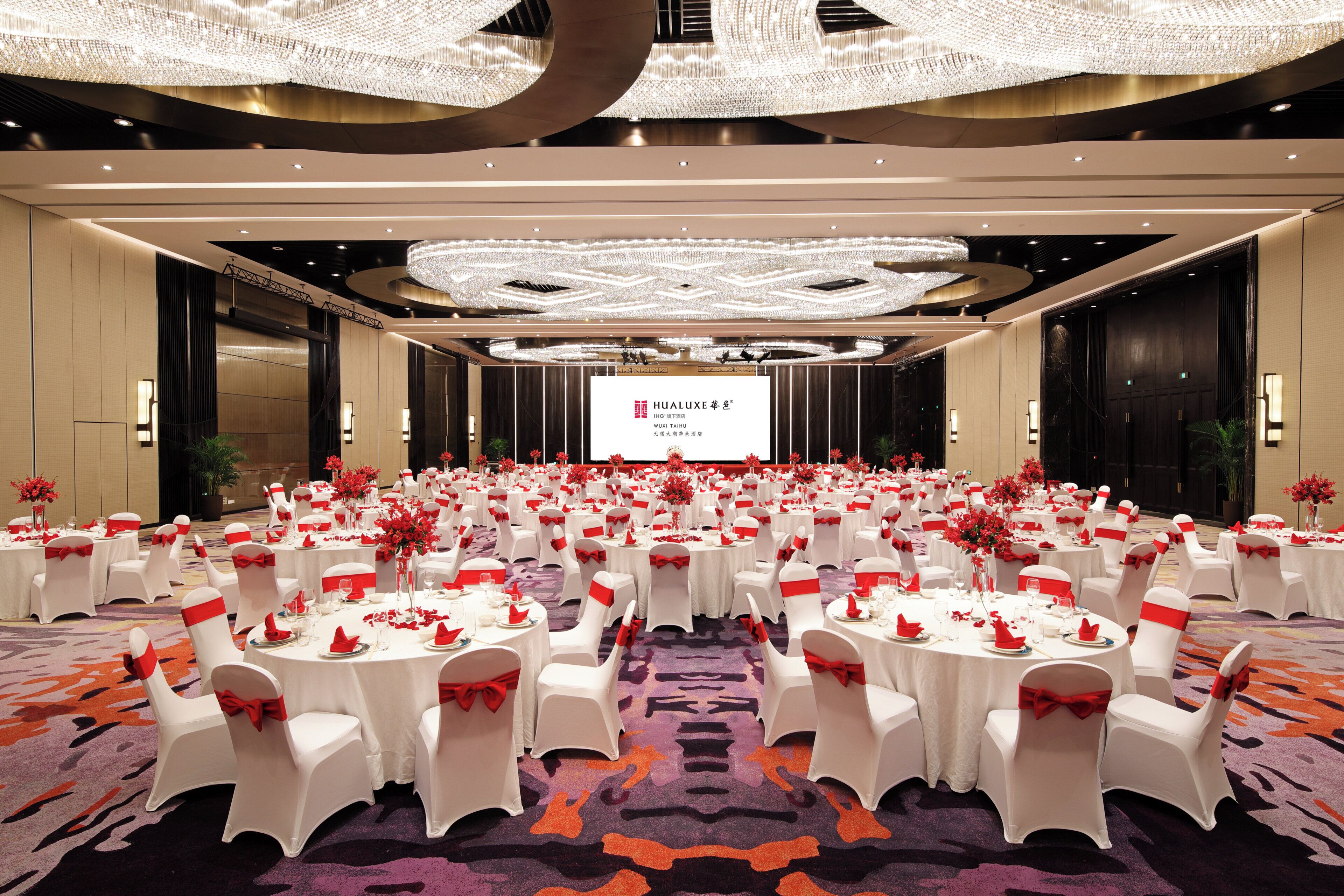 A large conference room with round tables, white chairs with red sashes, and a large screen at the front.