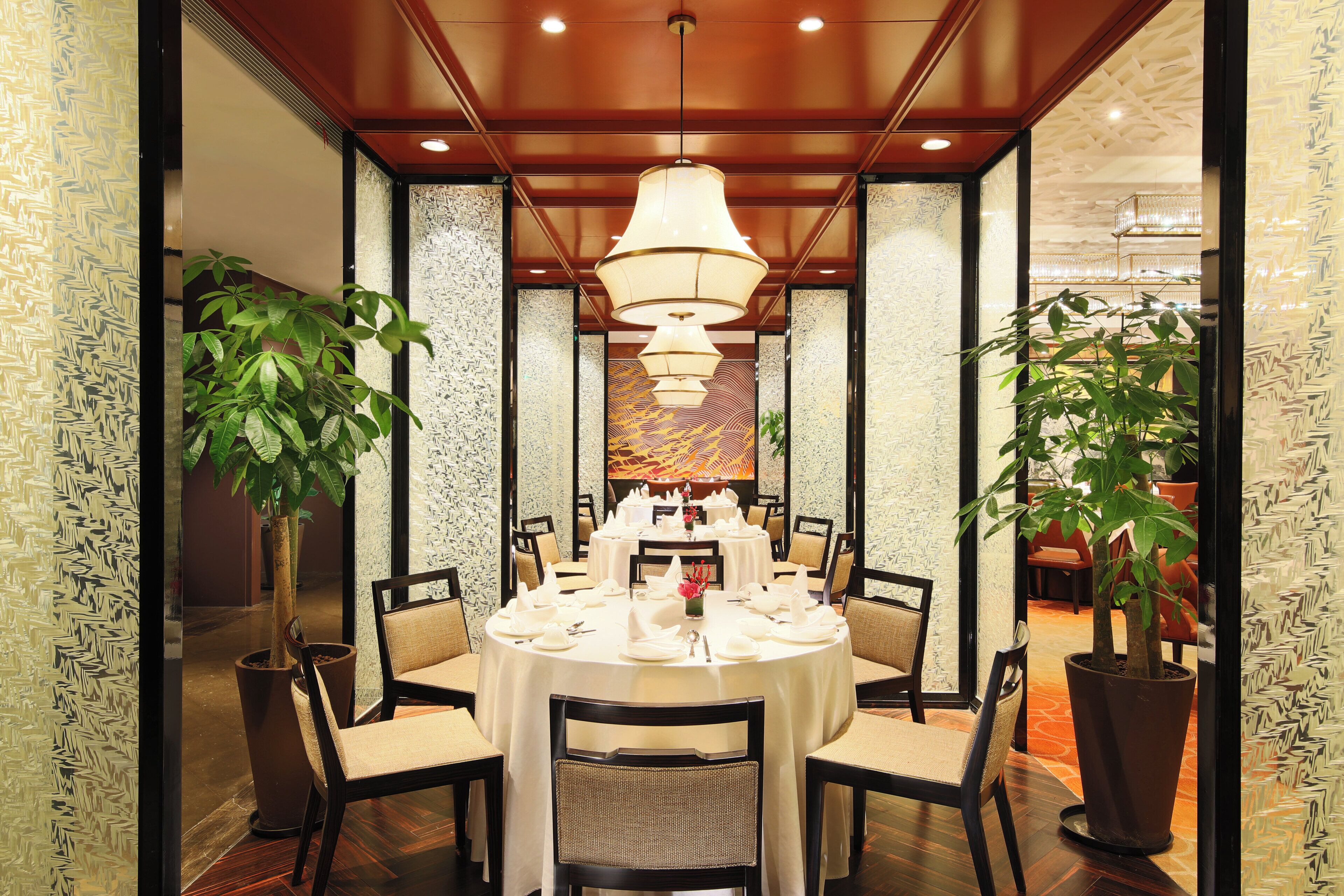 A dining area with round tables set for a meal, surrounded by chairs with woven seats, and large potted plants.