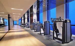 Un gimnasio moderno con equipo de ejercicio diverso, suelo de madera y una pared azul con un amplio ventanal.