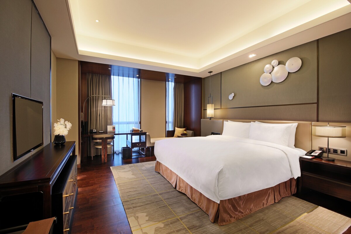 A hotel room with a large bed, a desk, a television, and a wall-mounted lamp.