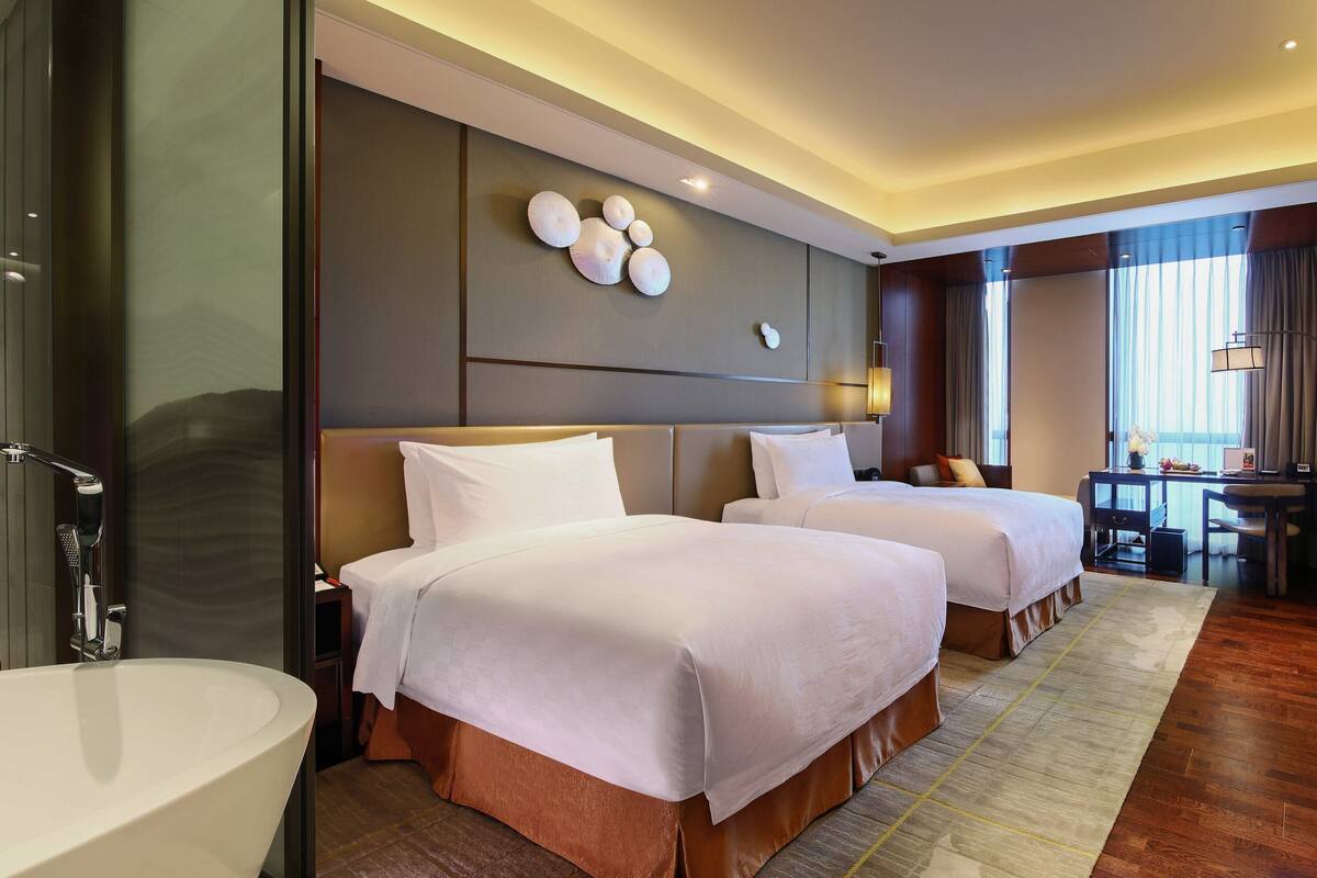 Ein Hotelzimmer mit zwei Betten, einem geräumigen Badezimmer mit Badewanne, einem Schreibtisch mit Stuhl und einem Fernseher.