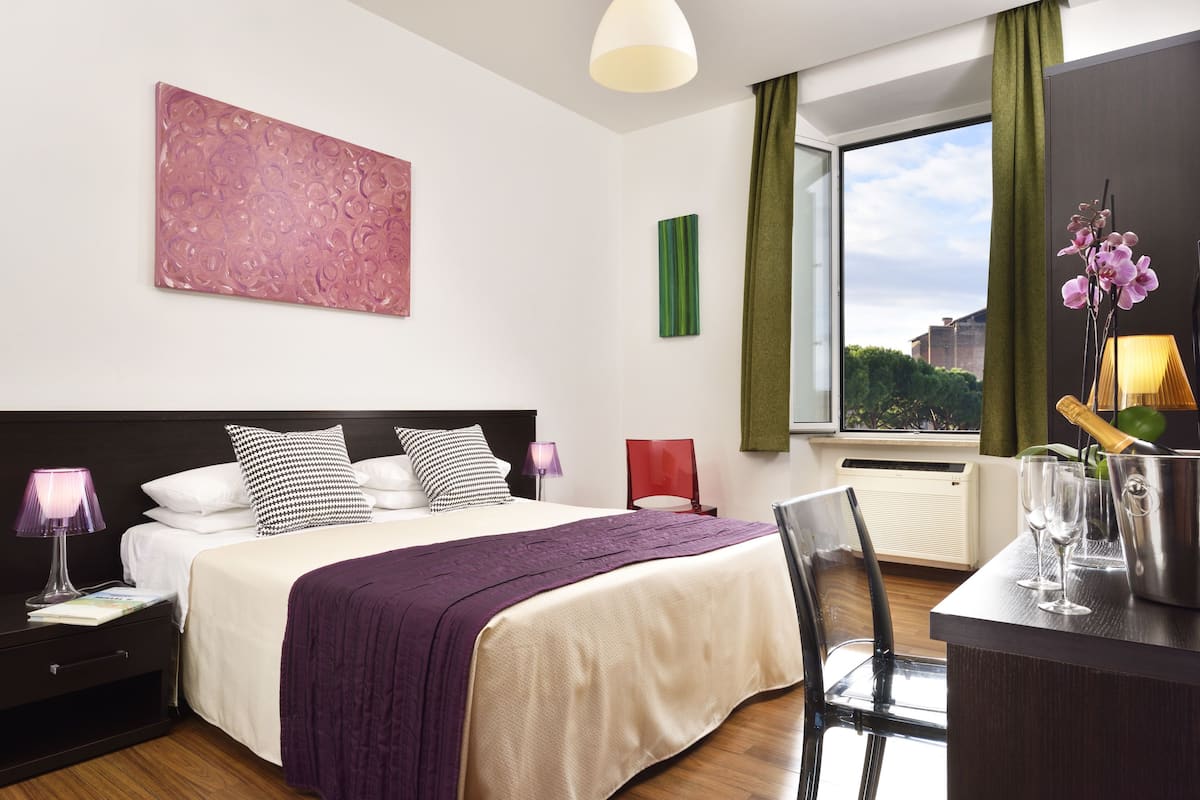 A modern hotel room with a large bed, a desk with a chair, a window with a view, and a green curtain.