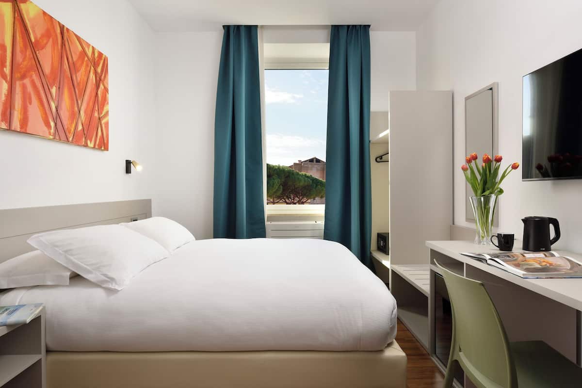 A hotel room with a bed, a desk, a chair, a window with curtains, a painting on the wall, and a vase with flowers.