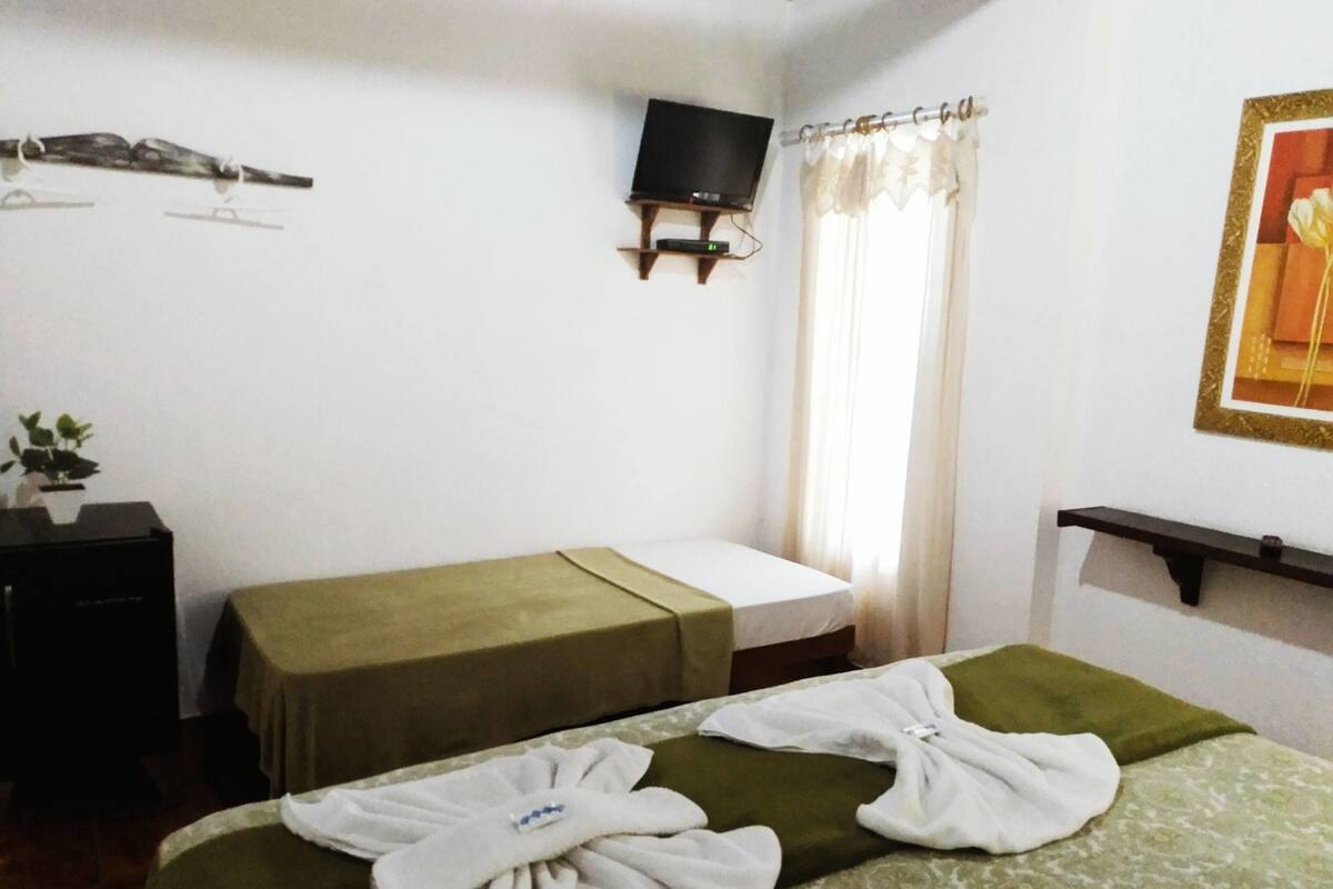 Ein Hotelzimmer mit einem Bett, einem kleinen Tisch, einem an der Wand befestigten Fernseher und einem Fenster mit Vorhängen.