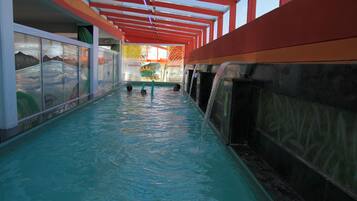 Piscina interna com água cristalina, uma parede de vidro de um lado e uma estrutura de teto vermelha e branca.
