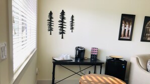 A small, cozy room with a window, a table with a coffee maker, a chair, and a decorative wall piece.