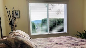 A bedroom with a bed, a window with blinds, a vase with branches, and a plant.