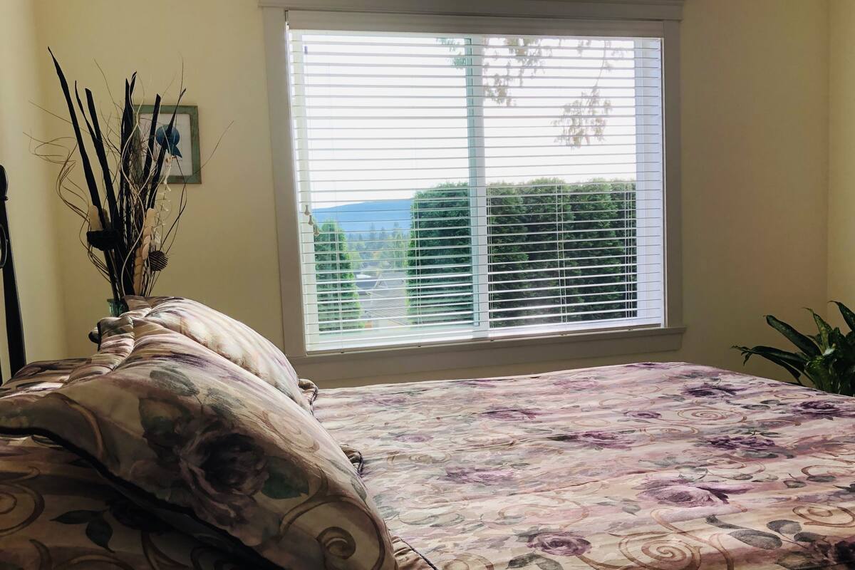A bedroom with a bed, a window with blinds, a vase with branches, and a plant.