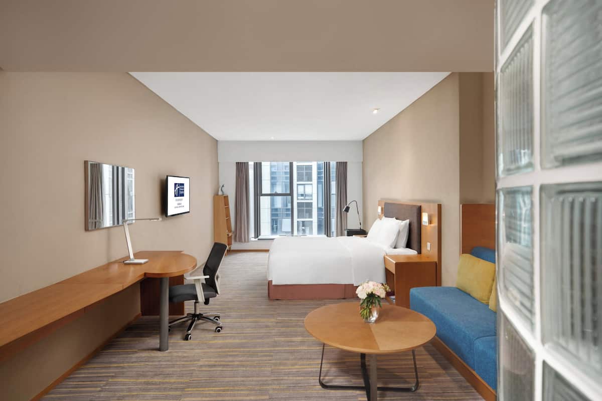 A hotel room with a bed, a desk, a chair, a small table with a vase, and a sofa.