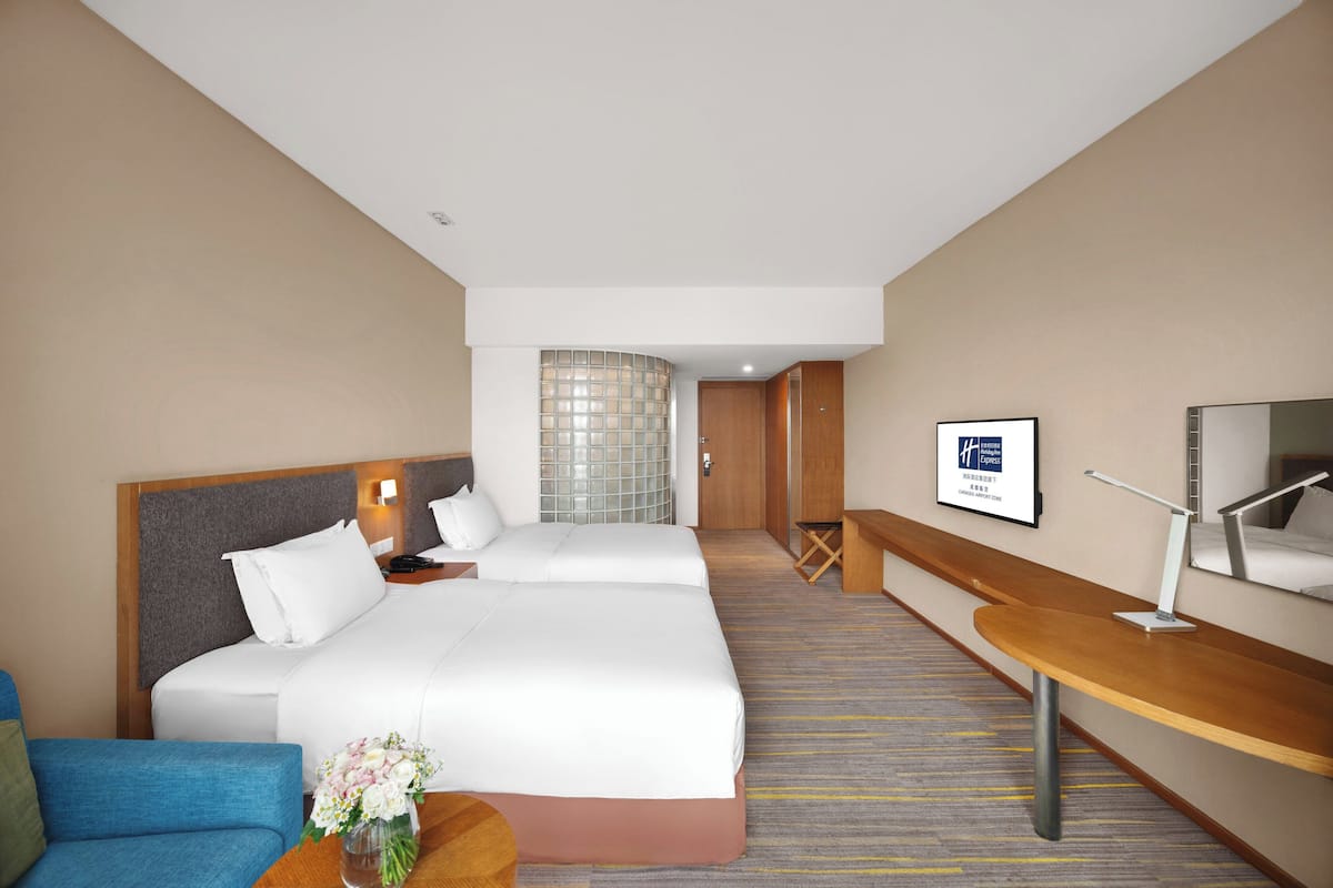 A hotel room with two beds, a desk, a chair, a small table with a vase, and a framed sign on the wall.