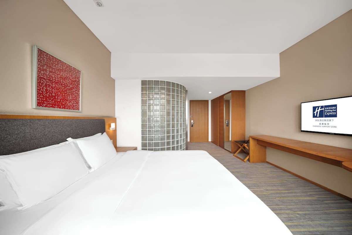 A hotel room with a large bed, a curved glass partition, a wooden bench, and a television on the wall.
