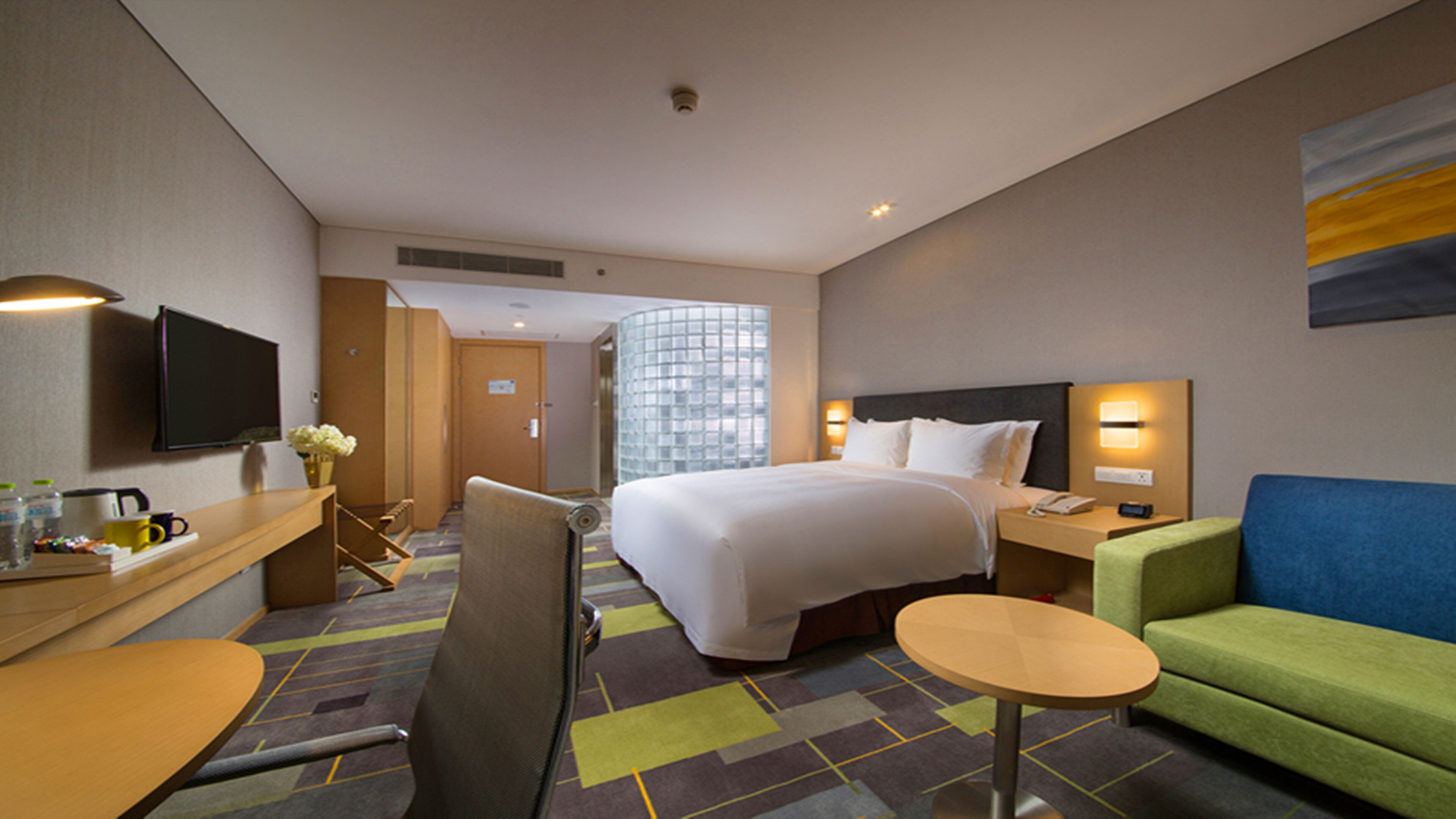 A hotel room with a bed, a desk, a chair, a small table, a sofa, and a TV.