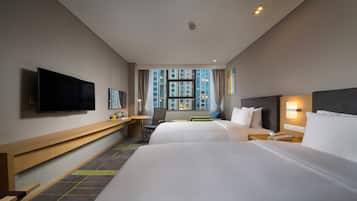 A hotel room with two beds, a desk, a flat-screen TV, a window with a city view, and a bedside lamp.