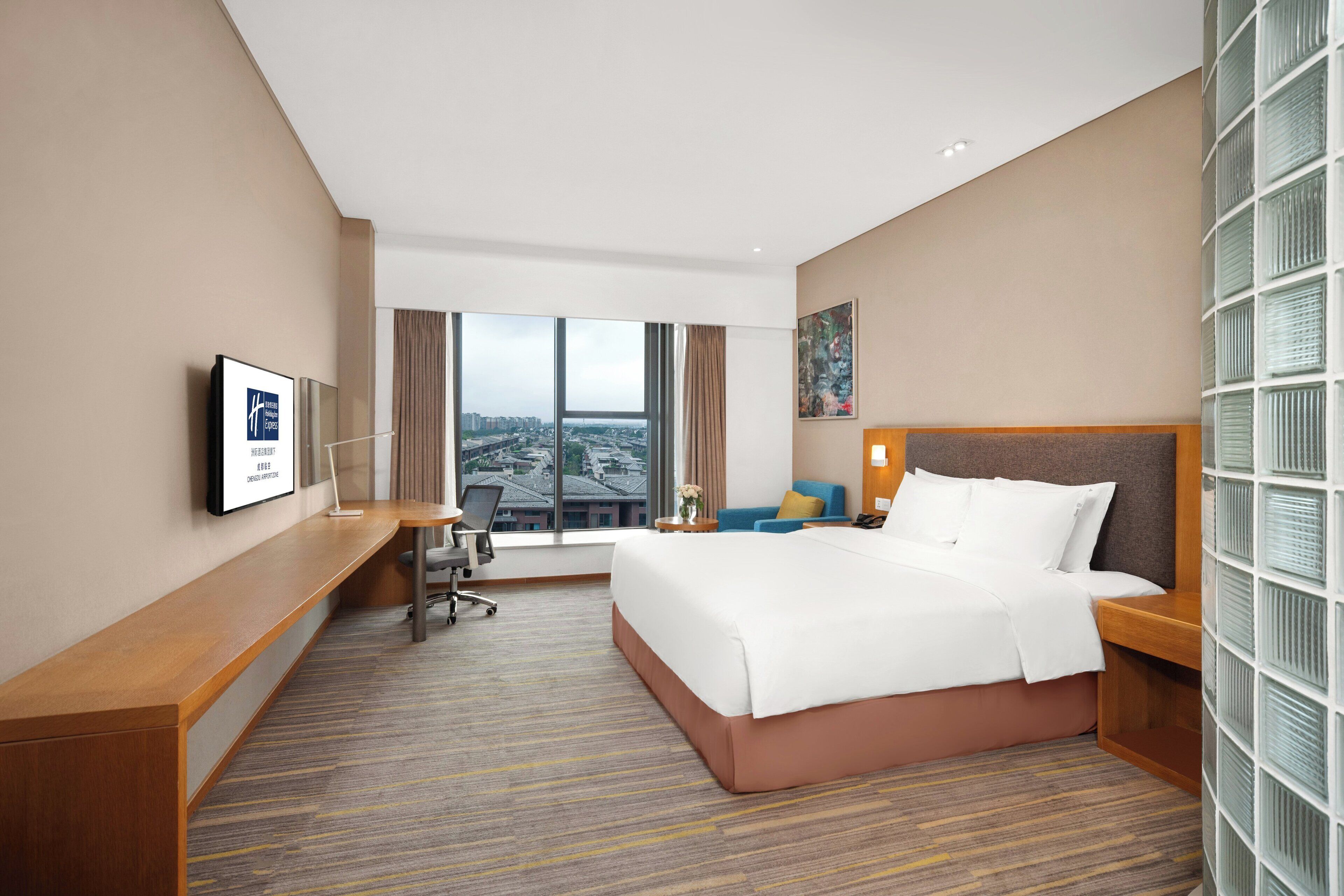 Habitación de hotel con una cama grande, un escritorio, una silla y vistas a la ciudad a través de amplias ventanas.
