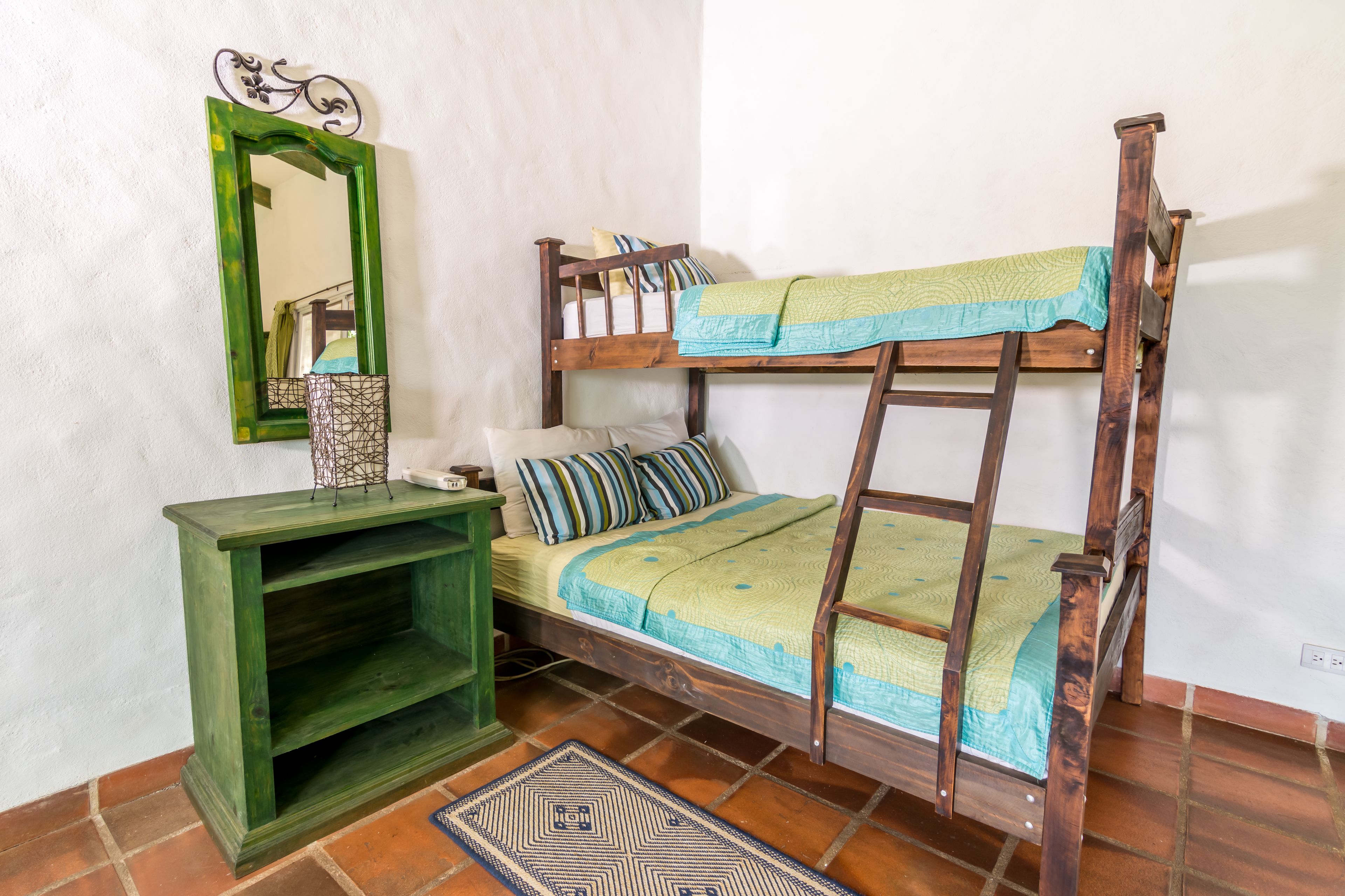 Una cama litera con una escalera, una mesita de noche verde, un espejo y una alfombra estampada en una habitación con paredes blancas y suelo de terracota.