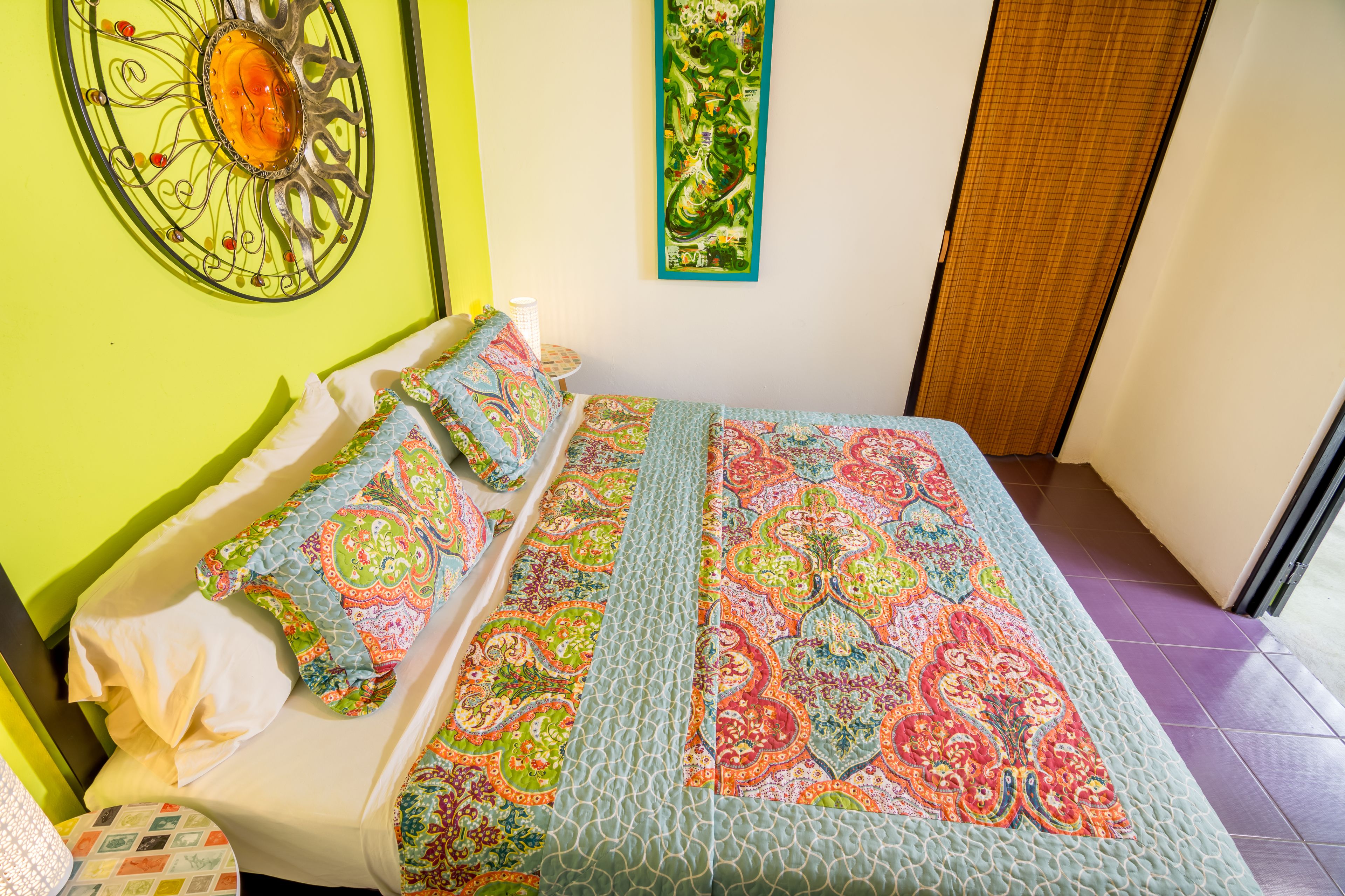 Una cama con un edredón colorido, un adorno de pared y una mesita con una lámpara.