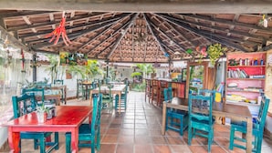 A covered outdoor dining area with wooden tables and chairs, surrounded by potted plants and a bookshelf. - Villa Margarita (La Garita)