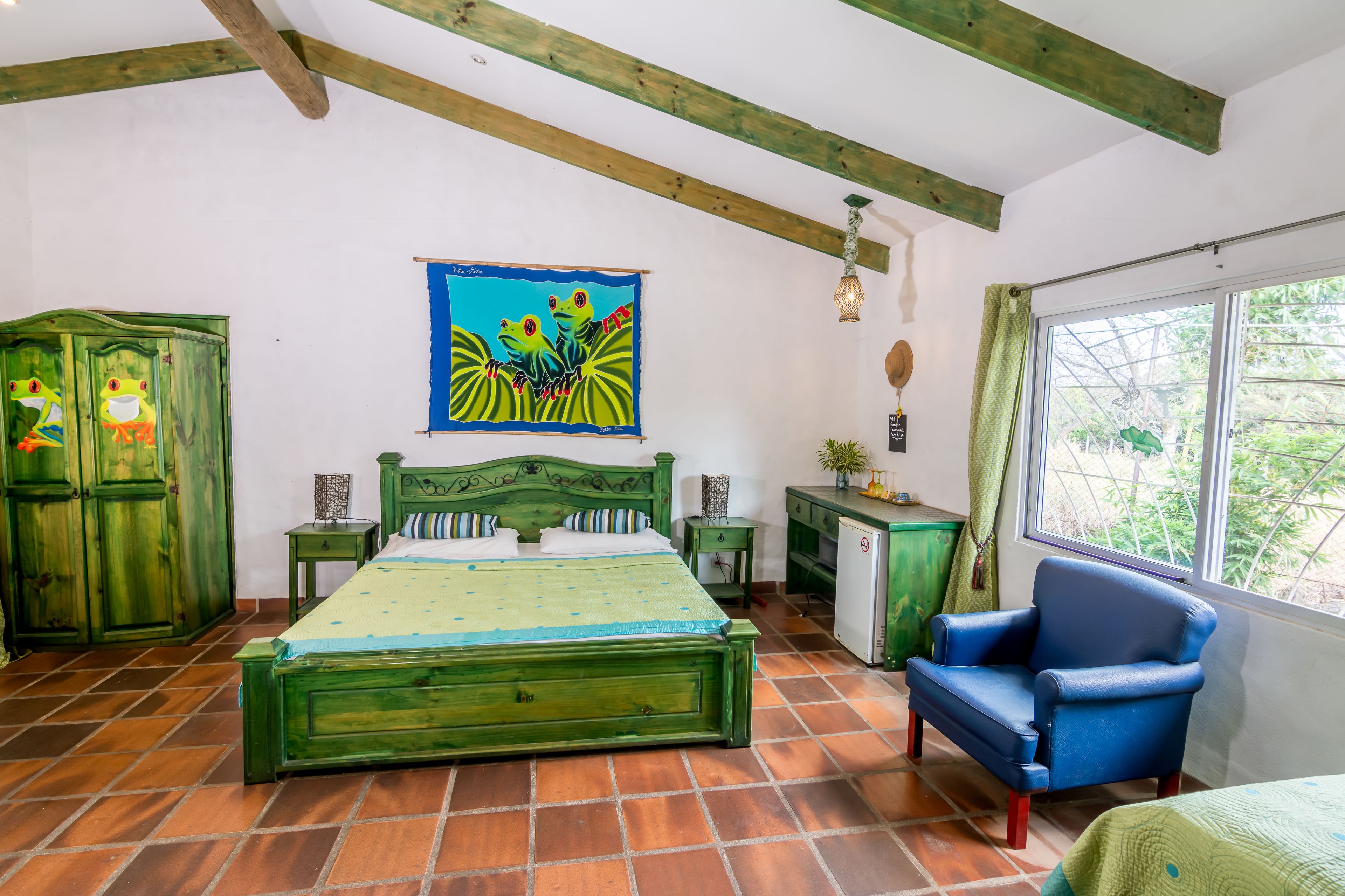 Een kamer met een groen bed, een blauwe stoel, een klein bureau en een raam met uitzicht op groen.