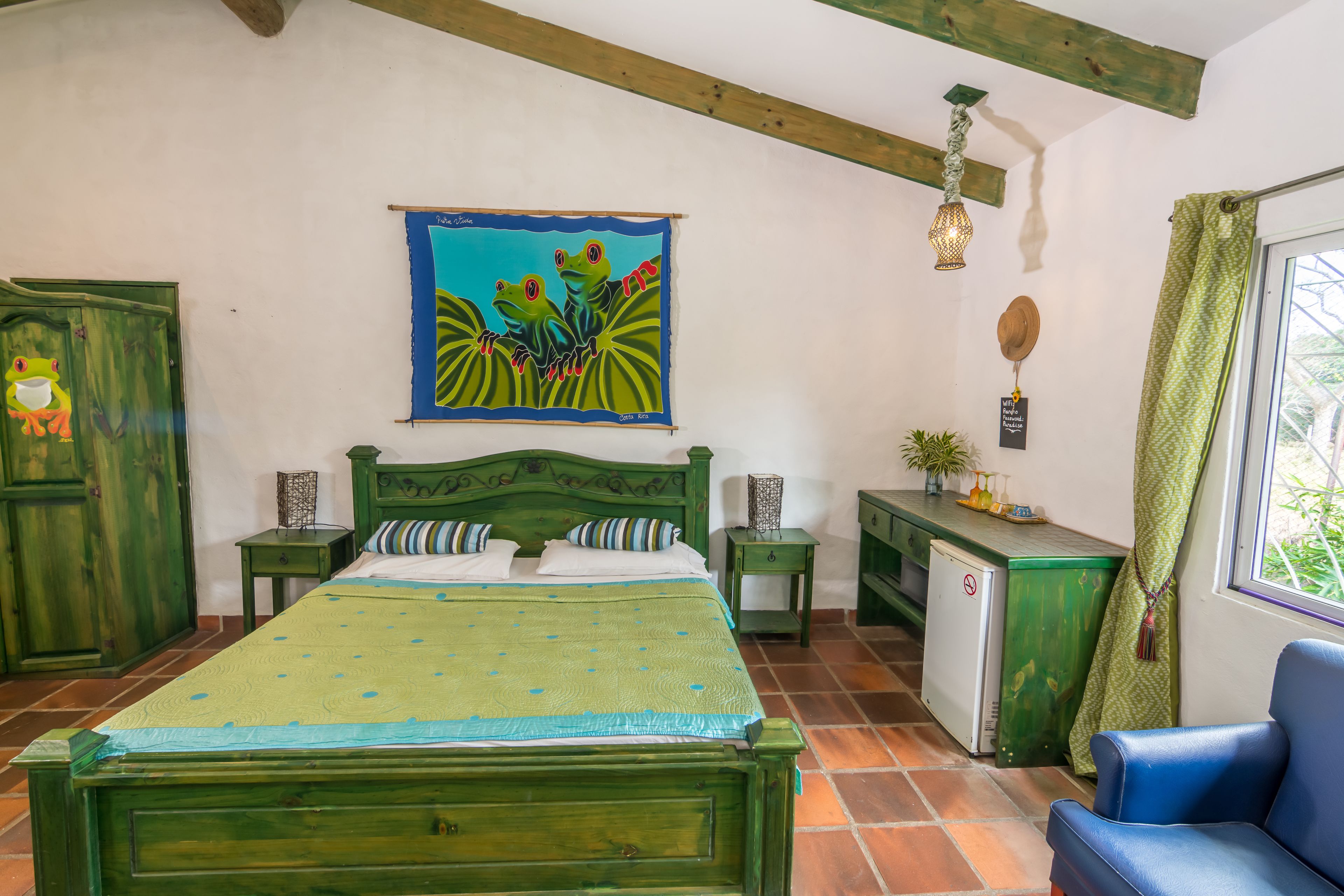 A room with a green bed, a small fridge, a desk, and a chair. There is a painting on the wall and a window with curtains.