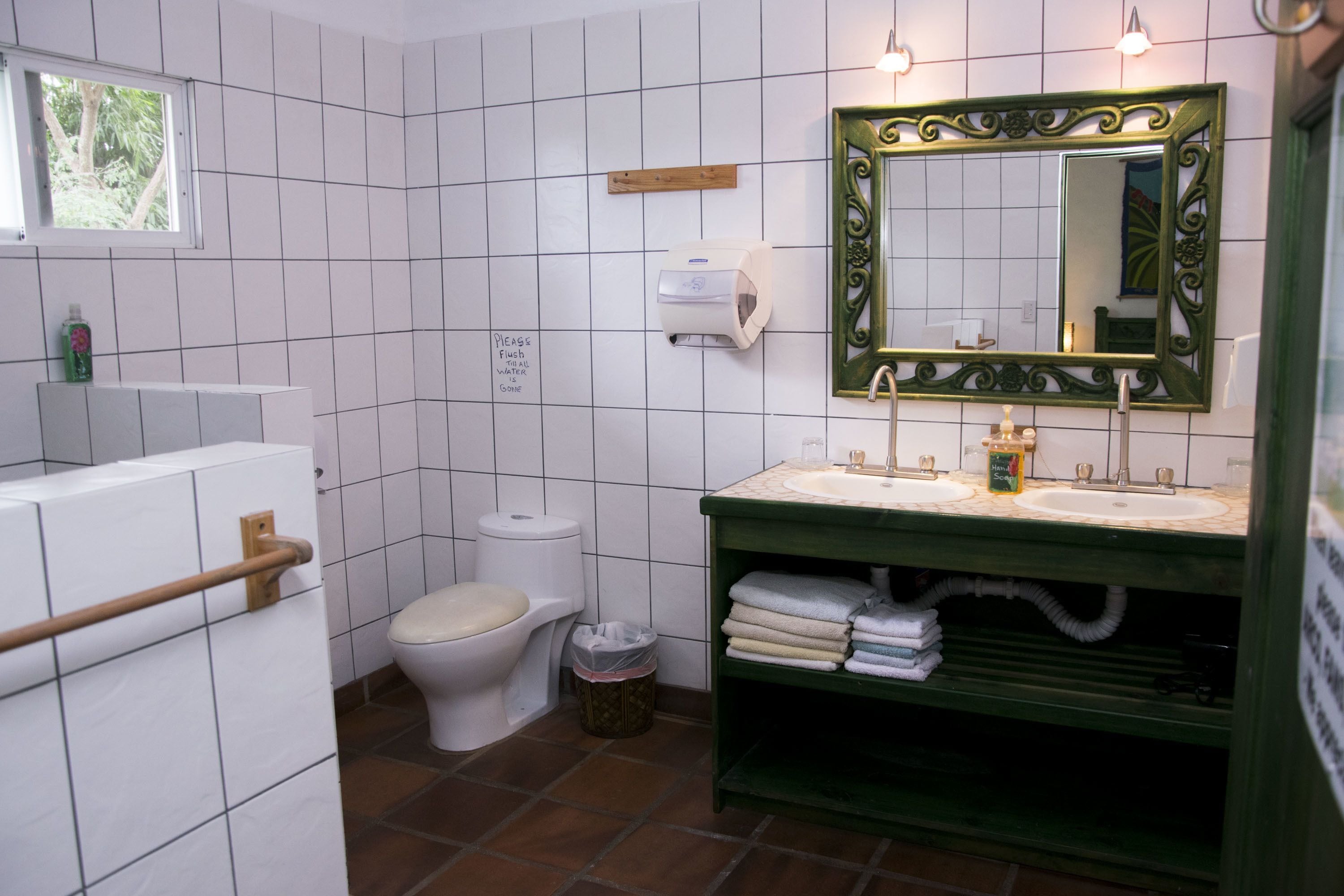 A bathroom with white tiled walls, a green vanity with two sinks, a toilet, and a hand dryer.