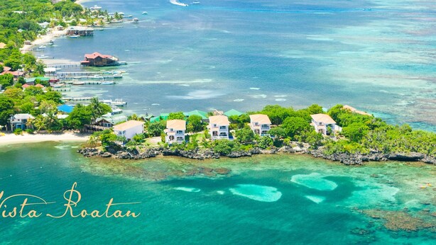 Vue aérienne de Coral Vista Roatan, avec un ensemble de bâtiments blancs aux toits rouges, entourés d’une végétation luxuriante et d’eaux turquoise cristallines.