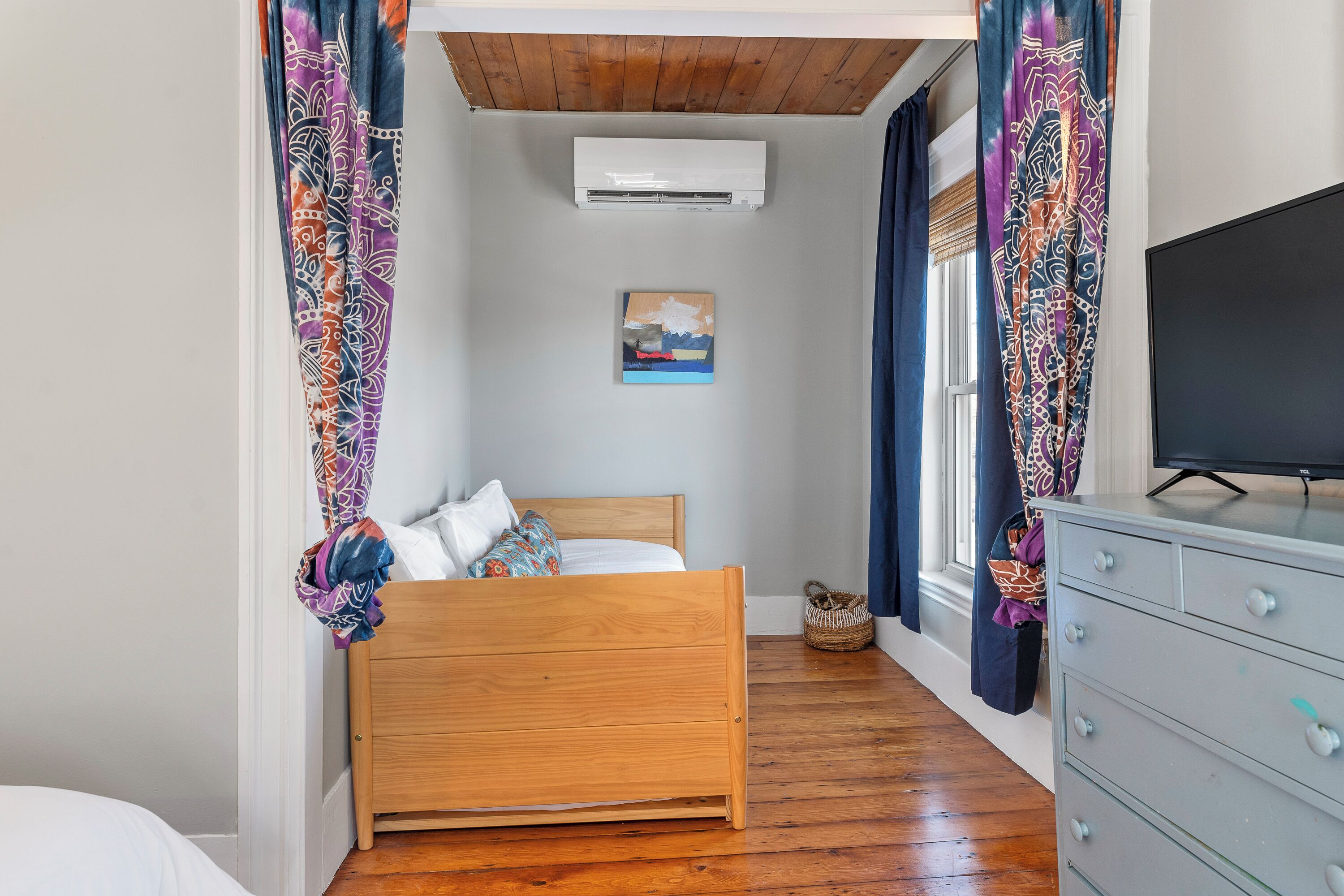 A bedroom with a wooden bed, an air conditioner, a television, and patterned curtains.