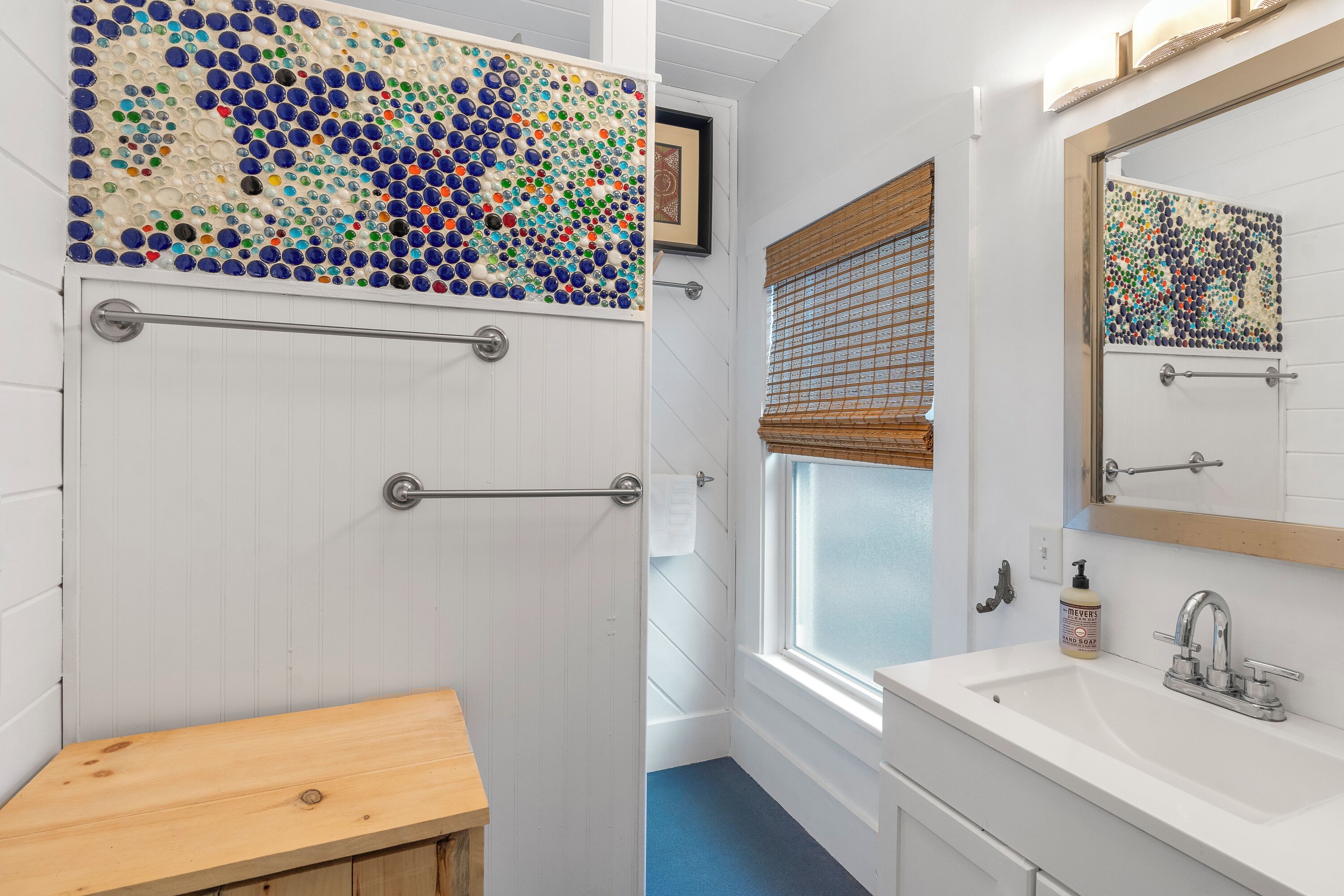 A bathroom with a mosaic wall, a wooden bench, a sink with a mirror, and a window with blinds.