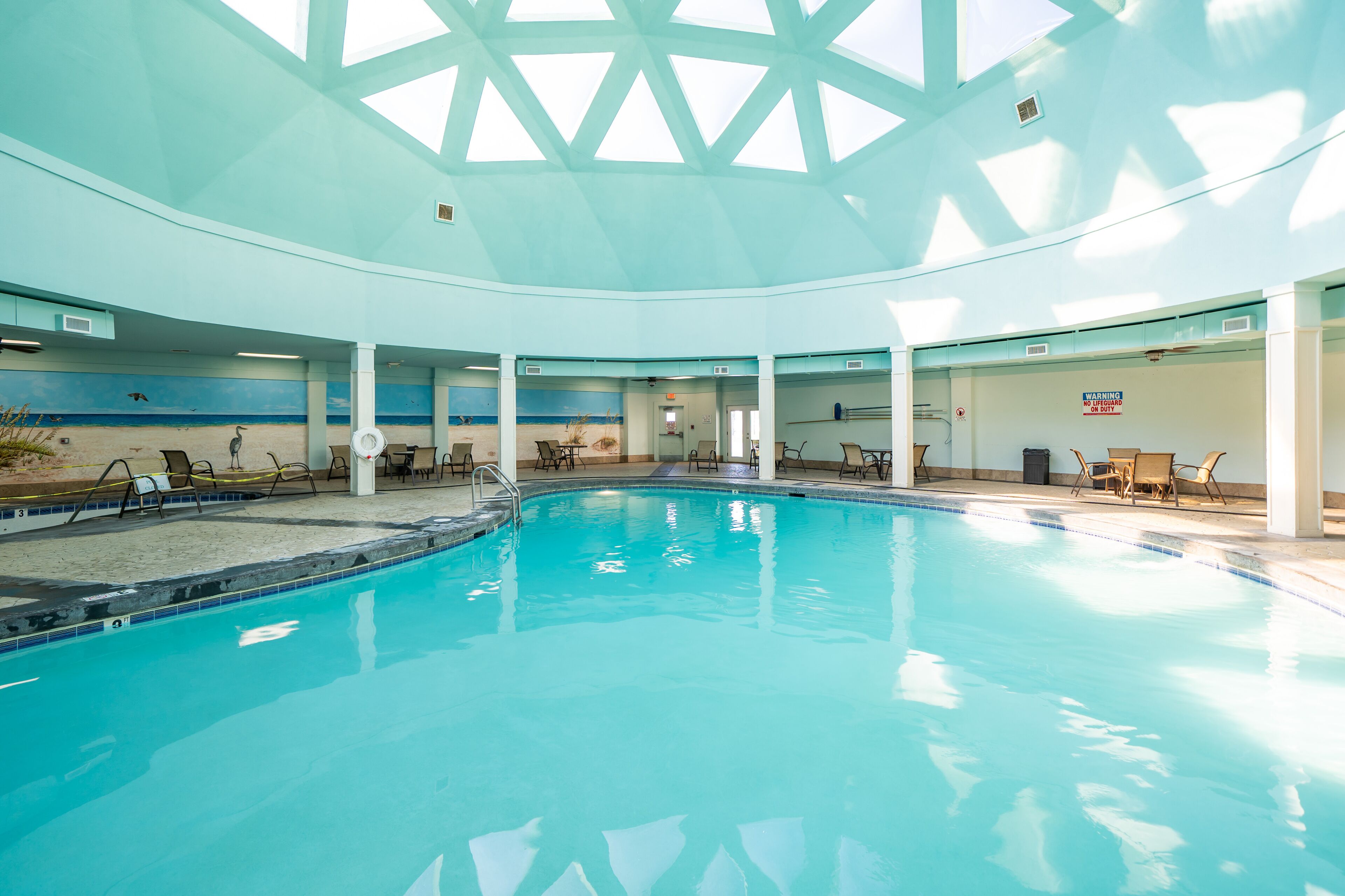 An indoor swimming pool with a high ceiling and skylights, surrounded by lounge chairs and a mural of a beach scene.