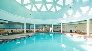 Una piscina interior con techo alto y tragaluces, rodeada de sillones de descanso y un mural con una playa.