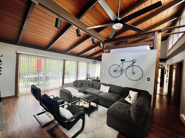 A spacious living room with a high ceiling and wooden beams, a large sofa set, a coffee table, and a bicycle wall art piece.