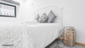 A neatly made bed with white bedding and decorative pillows, a wooden barrel by the wall, and a window with a metal grill. - Apartment with Patio Centro de Ovar (Ovar)