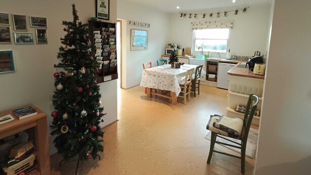 A cozy, well-lit room with a Christmas tree, a dining table with chairs, a kitchen area, and a wall with framed pictures.