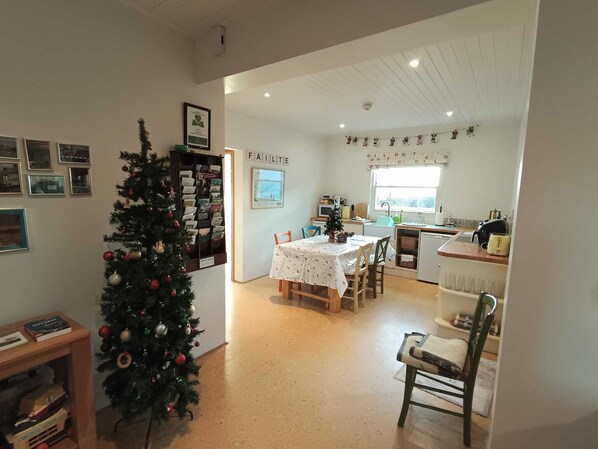 Een gezellige, goed verlichte kamer met een kerstboom, een eettafel met stoelen, een keukenhoek en een wand met ingelijste foto’s.