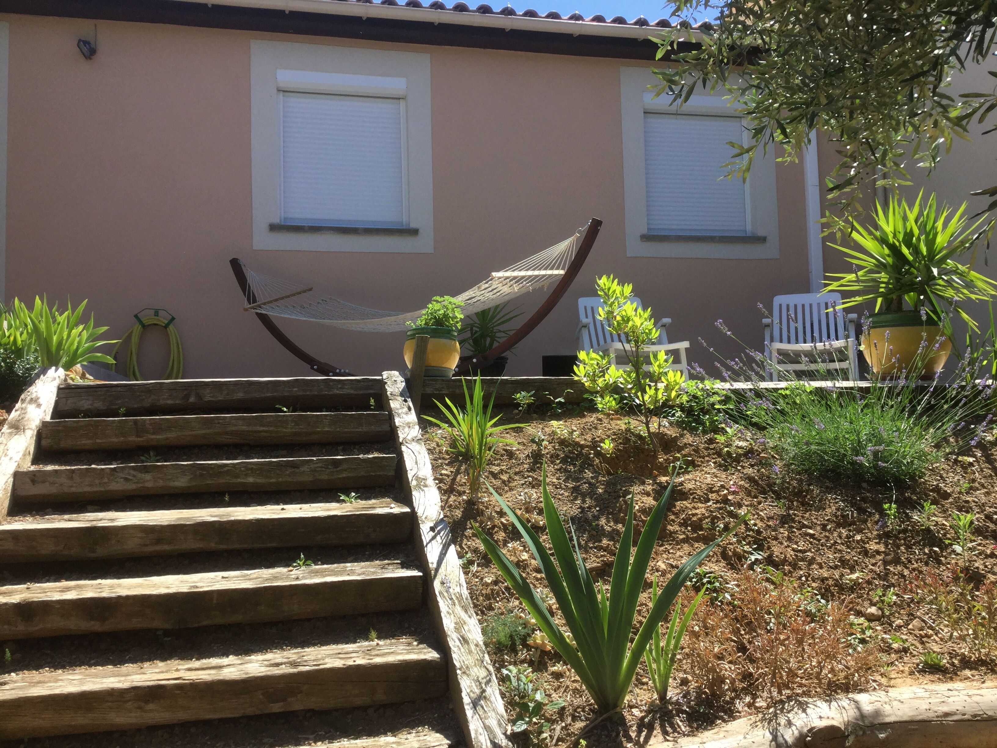 A wooden staircase leading up to a building with two windows, a hammock, and potted plants.