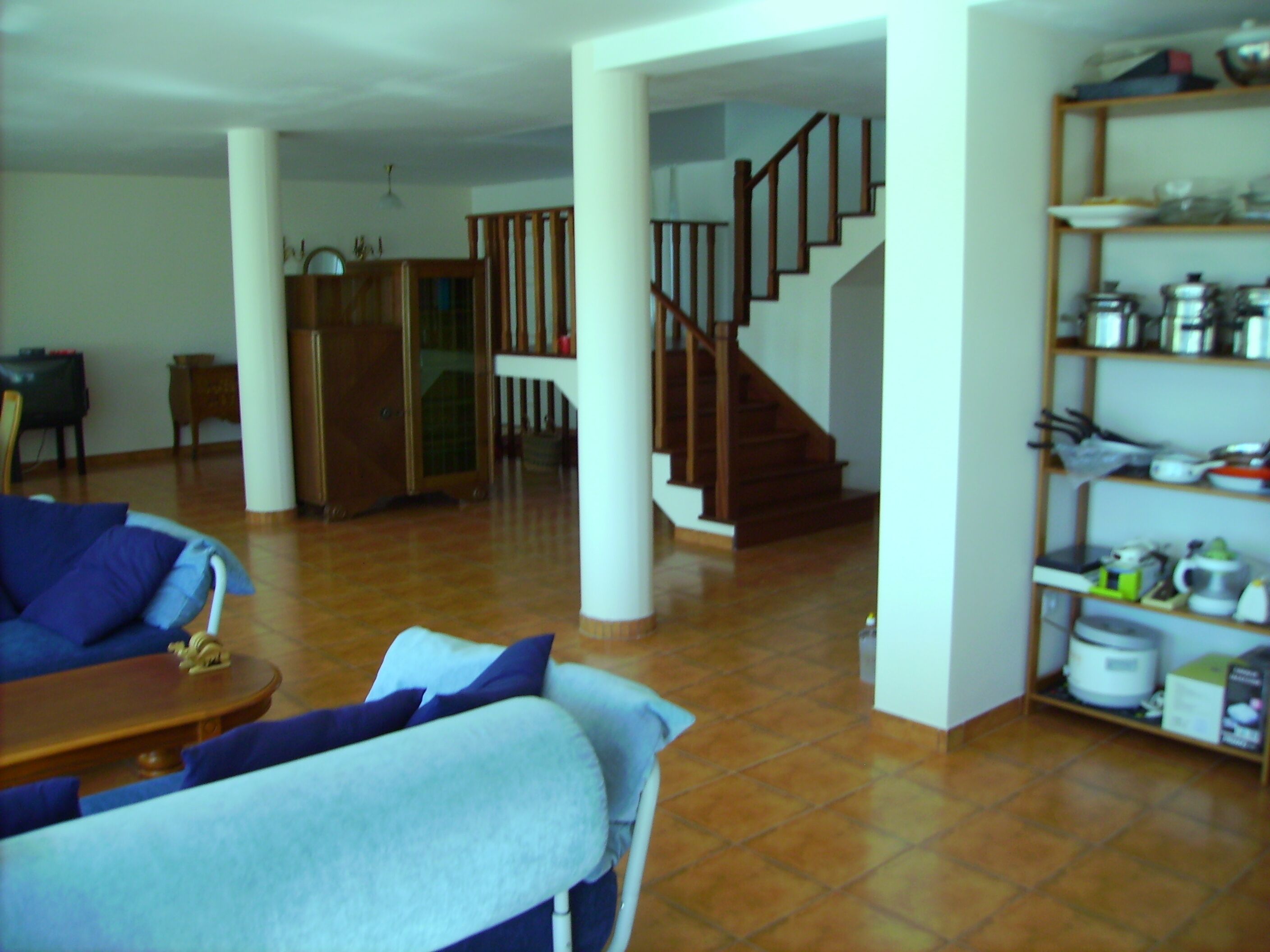 A spacious living area with a wooden staircase, a dining table, and a kitchen area with shelves.