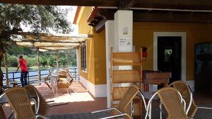 Ein Sitzbereich im Freien mit Korbstühlen und Tischen, einer Holzpergola und Blick auf ein Gewässer.