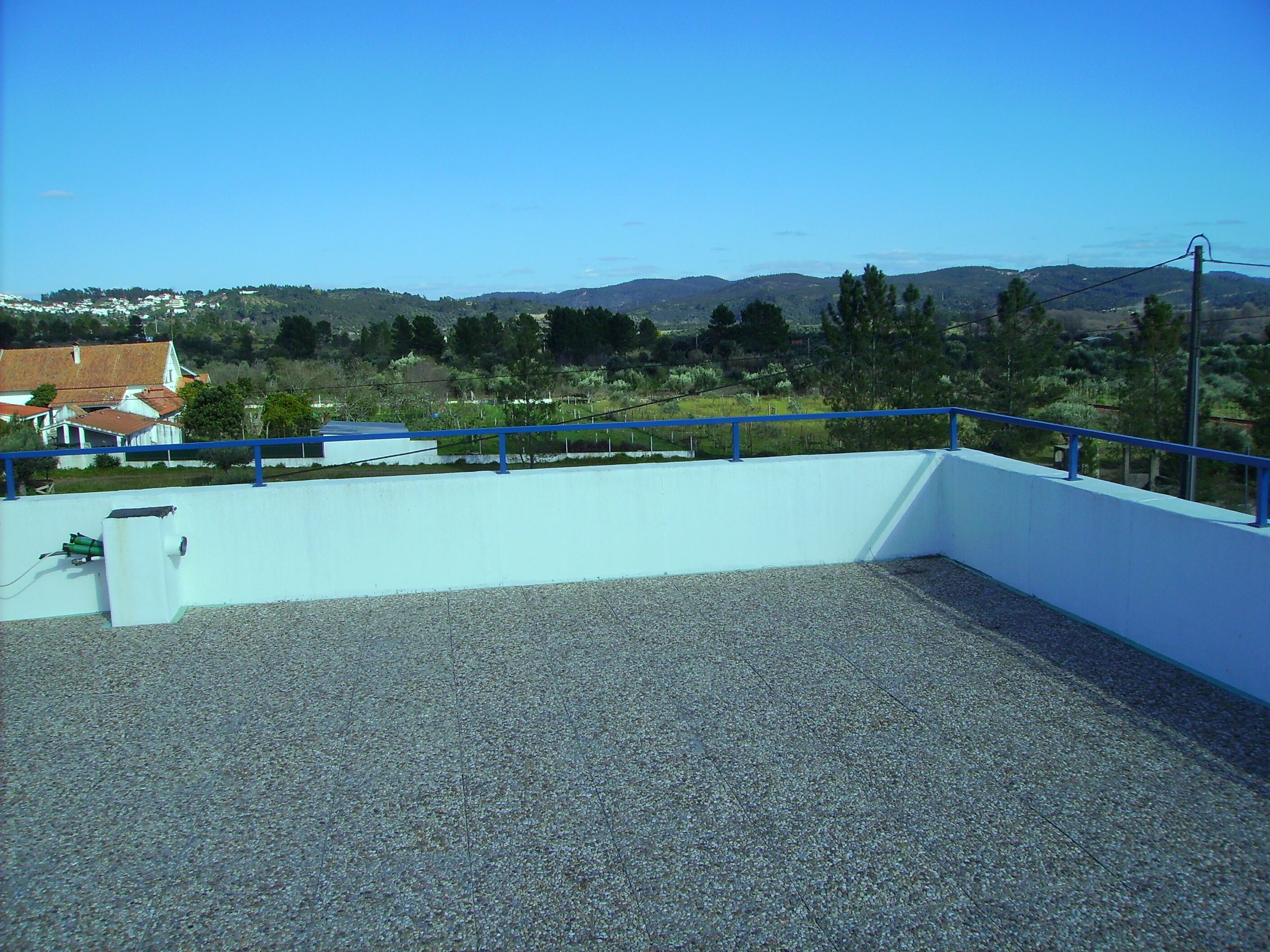 A rooftop terrace with a blue railing, gravel floor, and a view of a town with houses and hills in the background.