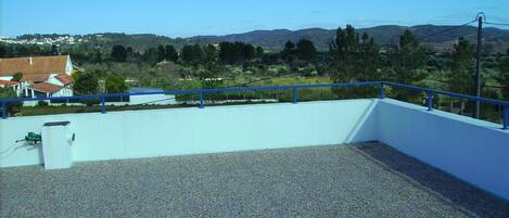 A rooftop terrace with a blue railing, gravel floor, and a view of a town with houses and hills in the background.