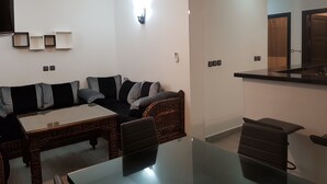 A modern living room with a glass-top dining table, a sofa with patterned cushions, a wooden coffee table, and a built-in kitchen area with a black countertop and wooden cabinets. - 3 modern new rooms with satelite wifi and air conditioning (agadir)