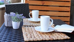 A wicker table with two white ceramic cups, a small potted plant, and a woven placemat on a balcony with a wooden bench and a palm tree in the background. - Arrive near the beach & feel good. (Sant Llorenç des Cardassar)