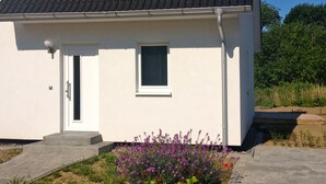 A white building with a dark tiled roof, a small window, and a door. There is a garden with purple flowers in front. - Beautiful, natural flat with big terrace in the heart of the island of Rügen (Patzig)