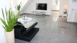 A modern living room with a black sofa, a round coffee table, a flat-screen TV mounted on the wall, and a potted plant. - Comf. mod. Apartment -Anker-(120m2) in the middle of Steinhude, wallbox, parking lot, WLAN, (Wunstorf)