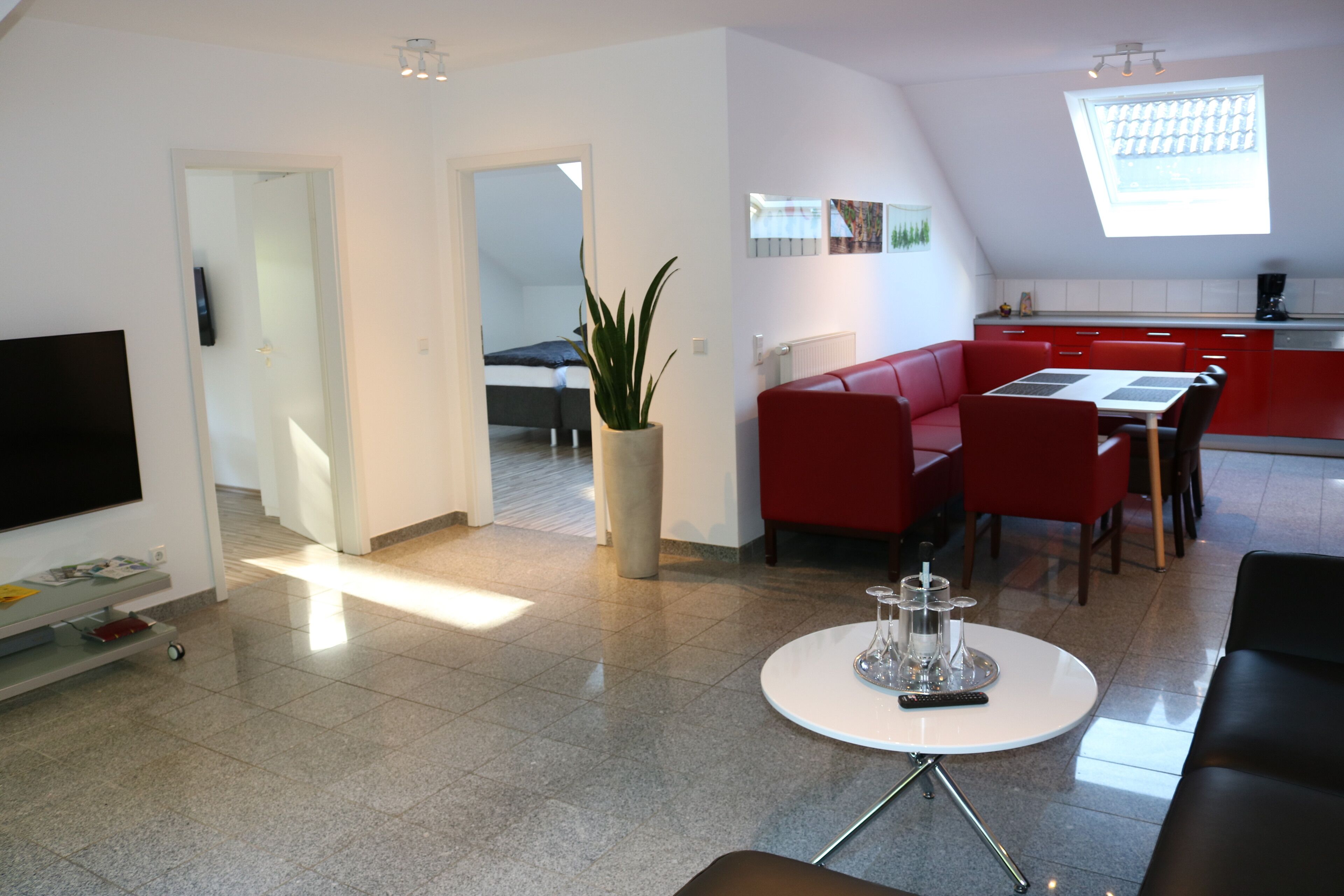A modern living room with a flat-screen TV, a dining area with red chairs, a white coffee table, and a kitchen in the background.