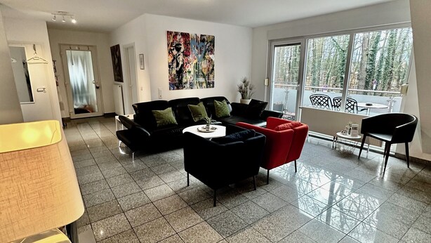 A spacious living area with a black sofa, a red armchair, and a coffee table. There is a large window with a view of trees and a balcony. - Comf. mod. Apartment -Anker-(120m2) in the middle of Steinhude, wallbox, parking lot, WLAN, (Wunstorf)