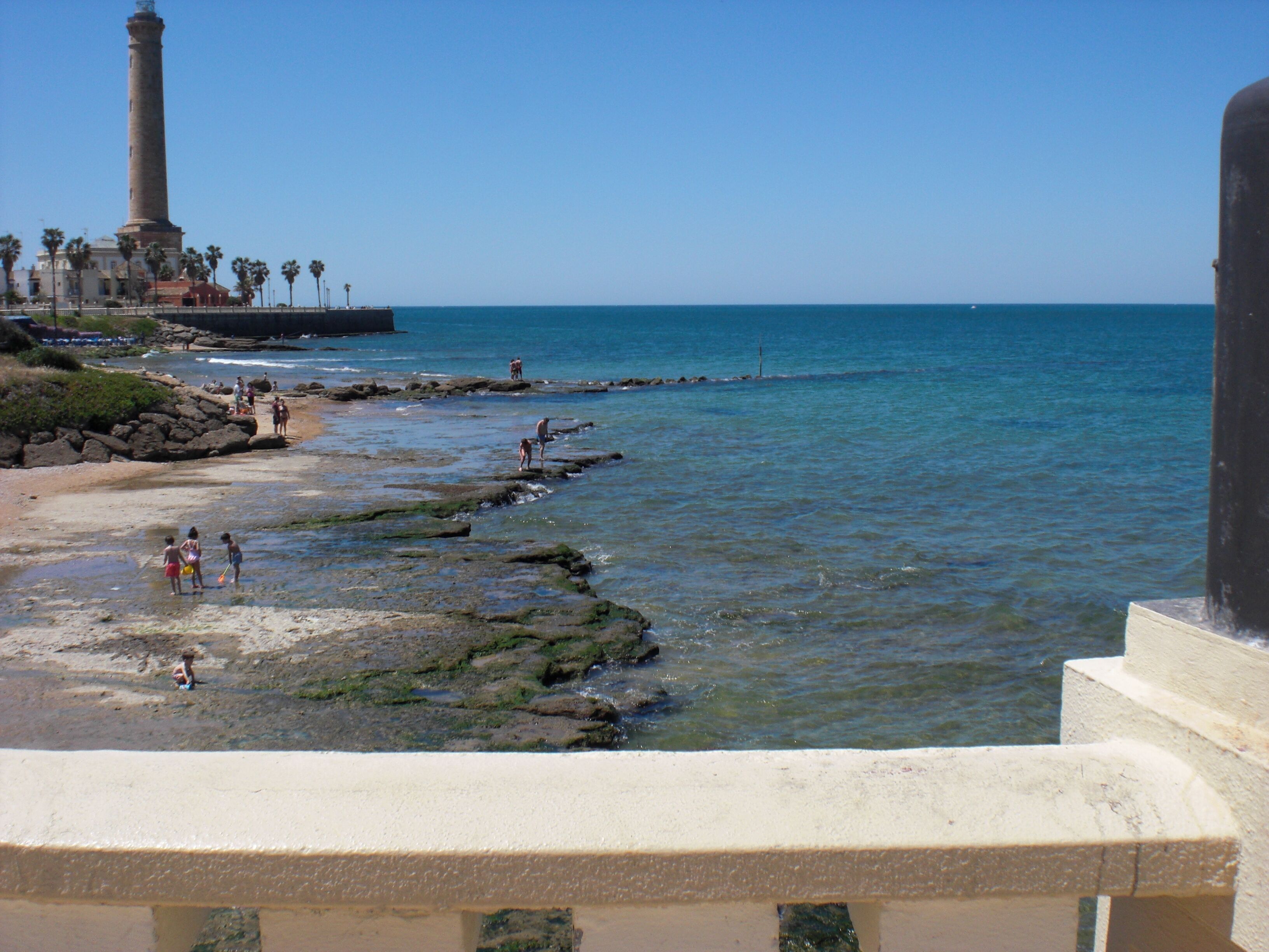 Vista costera con un faro, costa rocosa y mar azul claro.