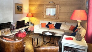 A cozy living room with a wooden ceiling, a white sofa with orange and blue cushions, a brown leather armchair, a small round coffee table, and a white side table with a red book and a decorative lamp. - Chalet at the foot of the slopes and 5 minutes from shops and ski school (Vars)
