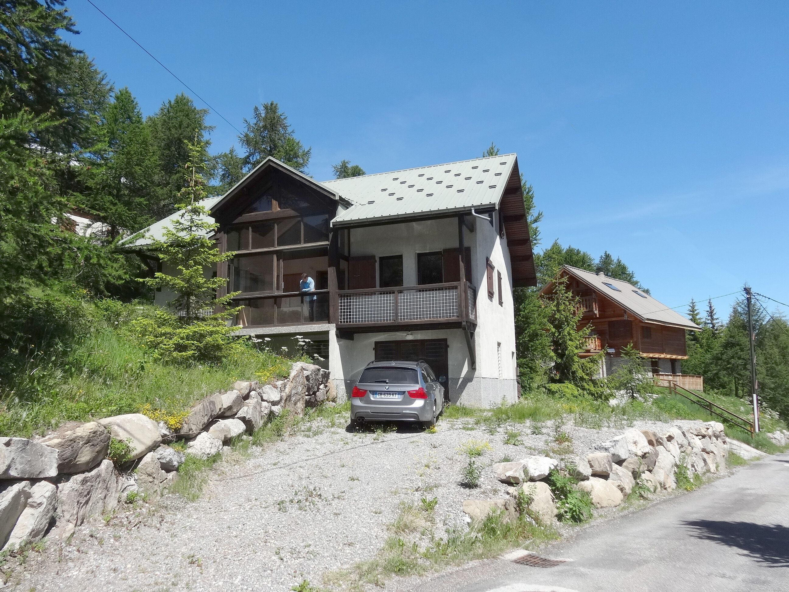 Un chalet à deux étages doté d’un balcon, d’un mur en pierre et d’une voiture garée.
