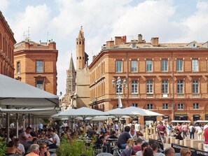 En livlig udendørs café med borde og parasoller foran historiske bygninger med røde murstensfacader og et markant tårn i baggrunden.