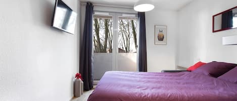 A bedroom with a large bed, a flat-screen TV mounted on the wall, a ceiling light, and a window with curtains.