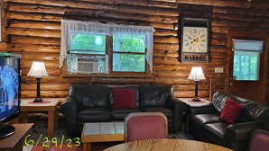 A cozy cabin interior with a wooden log wall, a leather sofa, a television, and a clock. - Stay WATERFRONT !   Stay CHEAT RIVER LODGE (Elkins)
